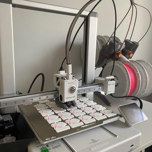 Peut inclure: Une imprimante 3D, étiquetée "Bambu Lab", est en fonctionnement, imprimant plusieurs objets blancs avec des motifs de rubans roses. L'imprimante est sur une surface blanche, avec une bobine de filament rose visible. La pièce est bien éclairée.