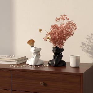 Puede incluir: Dos jarrones decorativos con forma de cabeza. Uno es blanco y contiene una brocha de maquillaje, el otro es negro y contiene flores rosas. Están sobre una cómoda de madera oscura con un collar de perlas y una vela blanca.