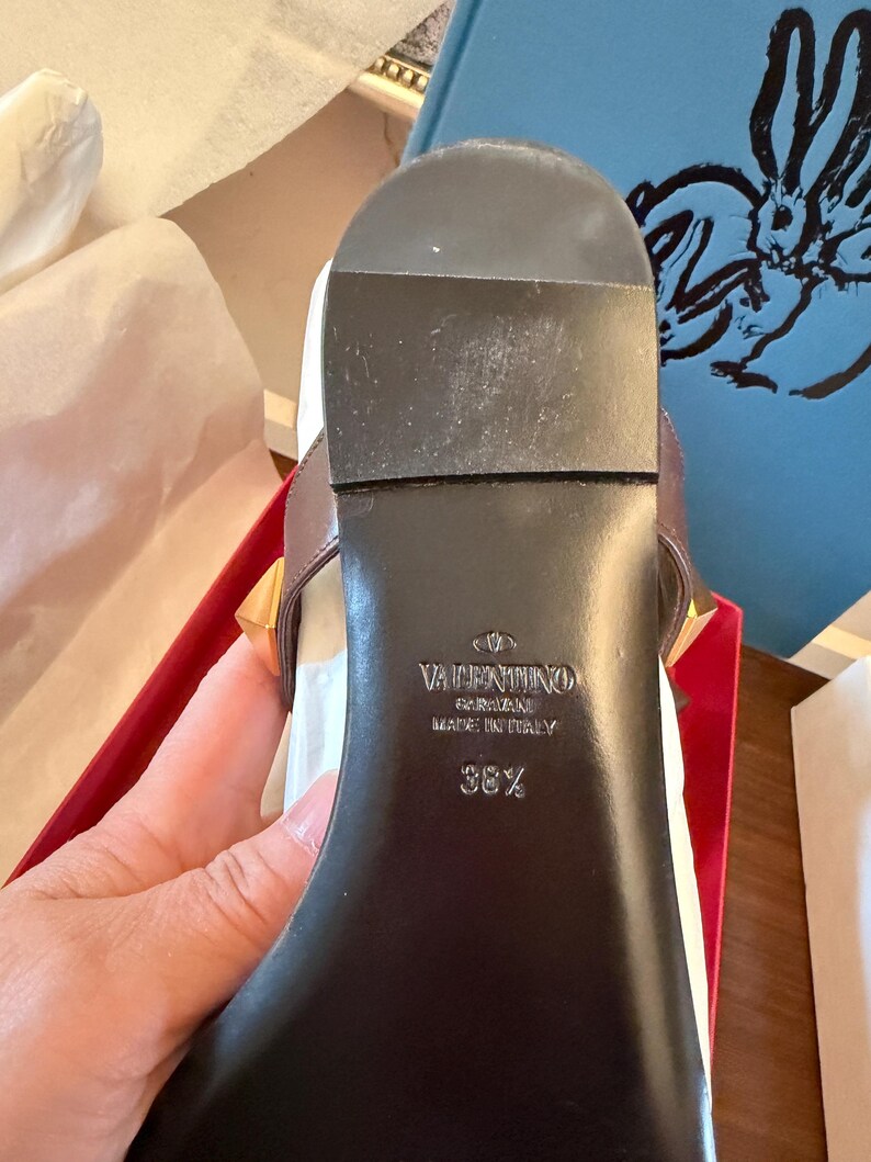 May include: Close-up of a brown leather sandal sole with gold-coloured studs. The brand name "Valentino Garavani" and "Made in Italy" are visible. The size is 38 1/2.
