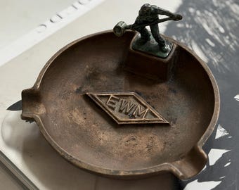 May include: A round, bronze-toned metal ashtray featuring a small, dark metal figurine of a person holding a hammer. The center of the ashtray displays a raised diamond-shaped emblem with the letters EWM embossed in the middle.