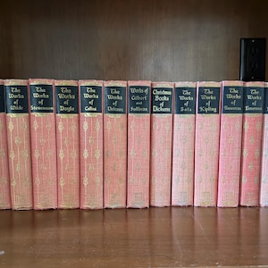 May include: A collection of antique books with pink covers and gold lettering, including titles like "The Works of Poe" and "Christmas Books of Dickens." The books are arranged on a wooden shelf, flanked by decorative bookends.
