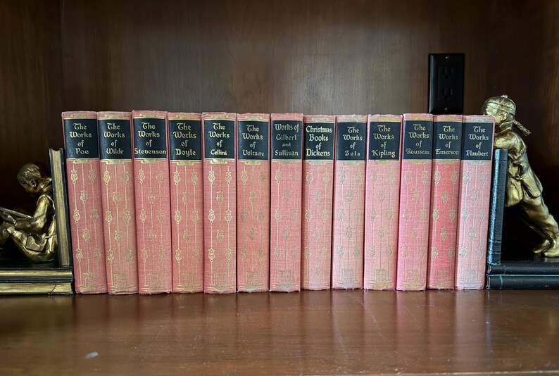 May include: A collection of antique books with pink covers and gold lettering, including titles like "The Works of Poe" and "Christmas Books of Dickens." The books are arranged on a wooden shelf, flanked by decorative bookends.