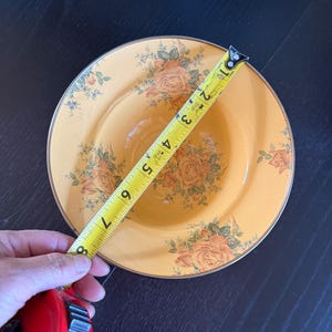May include: A round, yellow bowl featuring a floral pattern of orange roses and green leaves, with a gold-toned rim. A yellow measuring tape is placed across the bowl, indicating a diameter of approximately 8 inches from edge to edge.