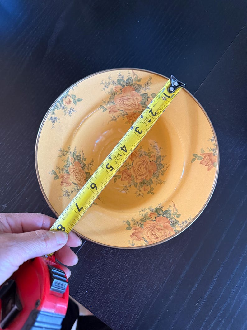 May include: A round, yellow bowl featuring a floral pattern of orange roses and green leaves, with a gold-toned rim. A yellow measuring tape is placed across the bowl, indicating a diameter of approximately 8 inches from edge to edge.