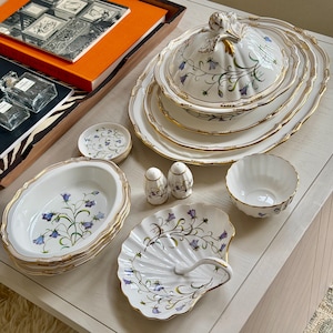 Op de afbeelding: Een verzameling witte porseleinen serveerschalen met gouden randen en bloemmotieven. De set bevat een serveerschaal met deksel, ovale schalen, kleine kommen, een schelpvormige schaal en zout- en pepervaatjes. Het bloemmotief heeft blauwe en groene accenten.