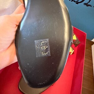 May include: Black leather sandal with gold and yellow details. The sole is black with a small symbol. The sandal is in a red box.