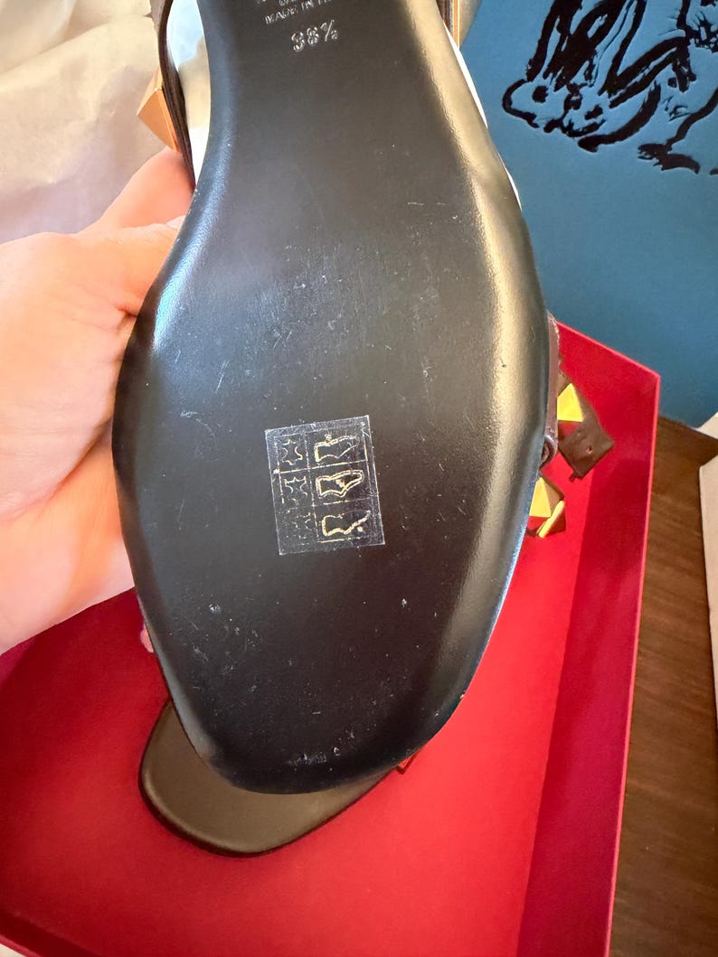 May include: Black leather sandal with gold and yellow details. The sole is black with a small symbol. The sandal is in a red box.