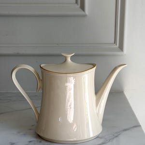 May include: A cream-colored ceramic teapot with a glossy finish, featuring a curved handle, a long tapered spout, and a matching lid with a small knob. The rim of the lid is accented with a thin gold-colored line, providing a subtle detail.
