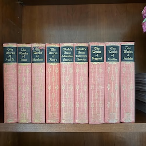 May include: A collection of antique books with pink covers and gold lettering. The titles include "The Works of Swift," "The Works of Ibsen," and "World's Great Adventure Stories." The books are arranged on a wooden shelf.