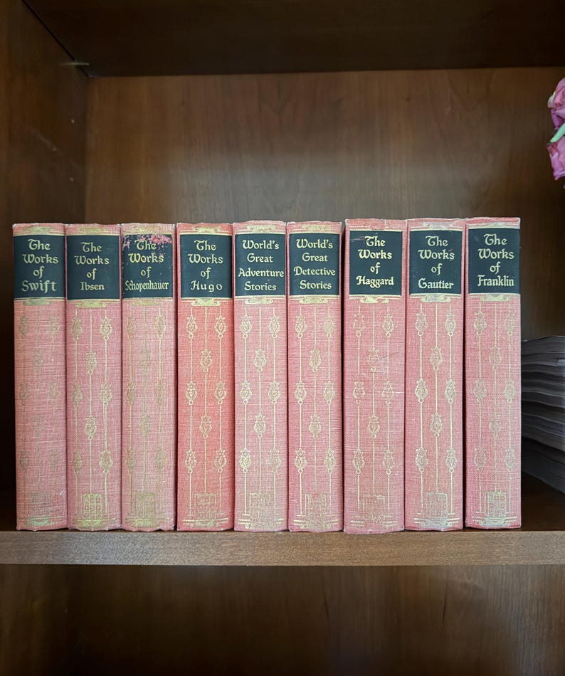May include: A collection of antique books with pink covers and gold lettering. The titles include "The Works of Swift," "The Works of Ibsen," and "World's Great Adventure Stories." The books are arranged on a wooden shelf.