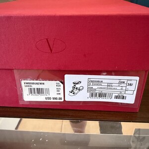May include: A red shoe box with a white label showing product details. The label includes a sandal illustration, size 38, and the price. The box features a large 'V' logo.