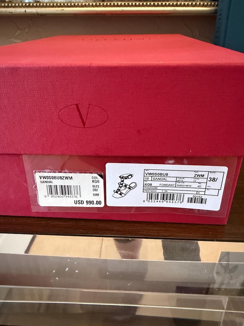 May include: A red shoe box with a white label showing product details. The label includes a sandal illustration, size 38, and the price. The box features a large 'V' logo.