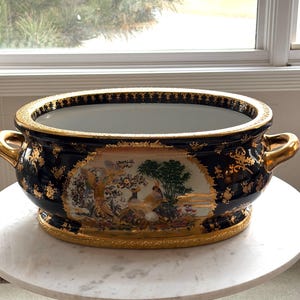 Op de afbeelding: Een ovale, zwarte keramische plantenbak met gouden accenten en handvatten. De plantenbak heeft een geschilderde scène met vogels en bomen, omlijst door gouden filigraan. De rand en de basis zijn ook afgewerkt met goud.