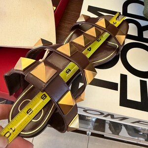 May include: Brown leather sandals with gold pyramid studs. The sandals have a flat sole and multiple straps. A measuring tape shows the sandals are approximately 25 cm long.
