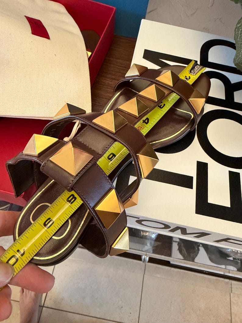 May include: Brown leather sandals with gold pyramid studs. The sandals have a flat sole and multiple straps. A measuring tape shows the sandals are approximately 25 cm long.