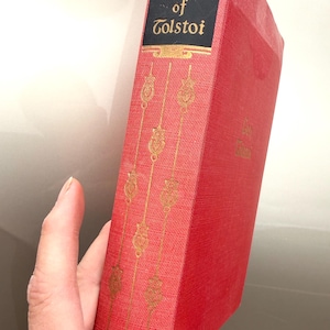 May include: A red book titled "The Works of Tolstoi" with gold lettering and decorative accents. The book's spine features the title on a black background. The cover has a textured appearance.