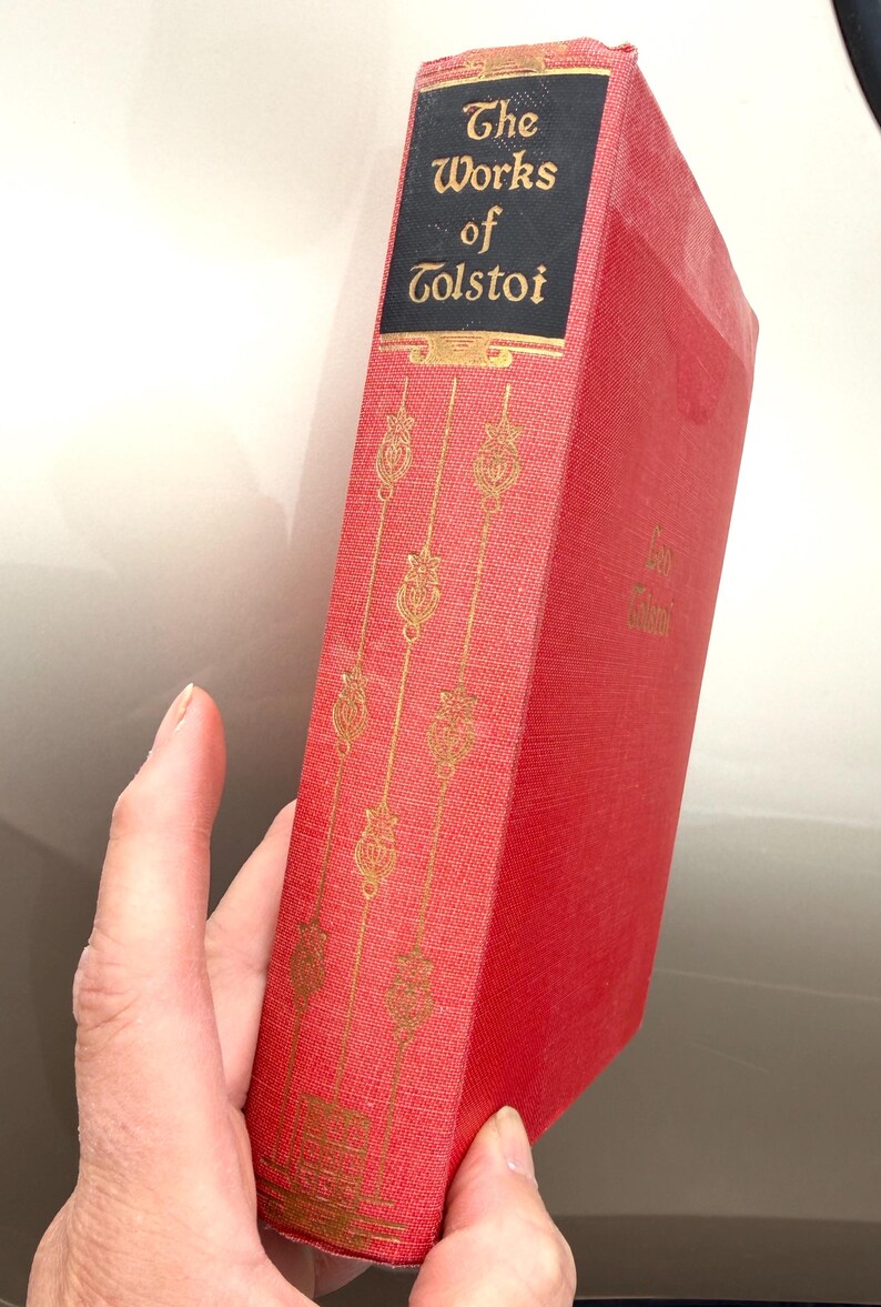 May include: A red book titled "The Works of Tolstoi" with gold lettering and decorative accents. The book's spine features the title on a black background. The cover has a textured appearance.