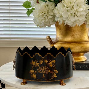 Puede incluir: Una jardinera ovalada de metal negro con borde superior festoneado, motivos de frutas y hojas pintados en oro, asas laterales con forma de cabeza de león y patas de garra. La jardinera tiene un acabado oscuro con detalles dorados.