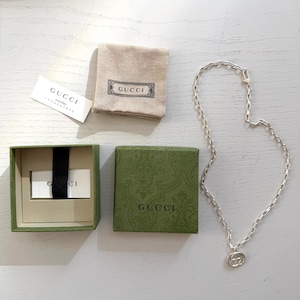 Puede incluir: Un collar de cadena de plata con un colgante circular que presenta el logotipo de Gucci, acompañado de una caja verde con una cinta negra, una bolsa de tela beige con una etiqueta Gucci y una tarjeta de autenticidad blanca Gucci controllato.