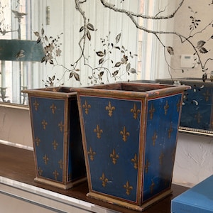 Puede incluir: Dos papeleras rectangulares de madera con acabado azul profundo y un patrón repetido de flor de lis dorada. Cada recipiente tiene una forma cónica, una base plana y bordes pintados con finas líneas rojas y doradas en la parte superior e inferior.
