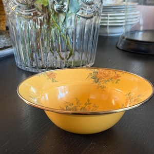May include: A round, yellow bowl with a metallic gold-toned rim. The interior of the bowl features a delicate floral pattern with orange roses and green leaves scattered across the surface. The bowl has a smooth, glossy finish and a wide, shallow shape.