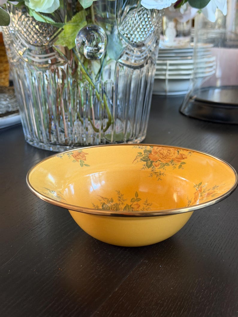 May include: A round, yellow bowl with a metallic gold-toned rim. The interior of the bowl features a delicate floral pattern with orange roses and green leaves scattered across the surface. The bowl has a smooth, glossy finish and a wide, shallow shape.