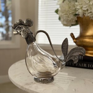 以下が含まれることがあります： 銀色の金属製の頭、取っ手、尾羽が付いた、ニワトリの形をした透明なガラス製デカンタ。デカンタは丸い大理石のテーブルの上に置かれています。背景には、窓と白い花が入った金の花瓶があります。