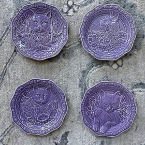 May include: Four round lavender ceramic plates with scalloped edges, each featuring a raised relief design of a cat surrounded by various flowers. The plates have a decorative lattice pattern along the rim and a glossy finish on the surface.