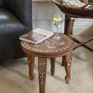 Puede incluir: Una mesa auxiliar redonda de madera con intrincados patrones de incrustaciones blancas en la superficie y las patas. La mesa tiene cuatro patas talladas y una parte superior circular decorada con motivos florales y geométricos. Un libro Indian Handicrafts.