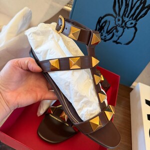 May include: Brown leather sandals with gold pyramid studs. The sandals feature multiple straps and a buckle fastening. They are presented in a red box with white tissue paper.