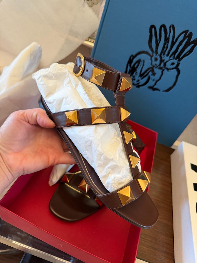 May include: Brown leather sandals with gold pyramid studs. The sandals feature multiple straps and a buckle fastening. They are presented in a red box with white tissue paper.