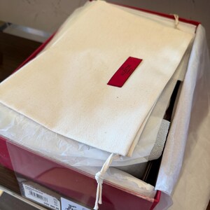 May include: A red box, partially open, containing a white fabric dust bag with a red Valentino label, tissue paper, and a product label. The contents are visible.