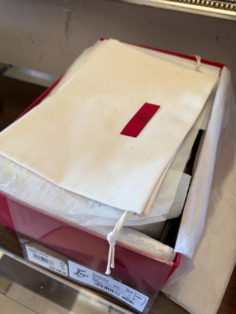 May include: A red box, partially open, containing a white fabric dust bag with a red Valentino label, tissue paper, and a product label. The contents are visible.