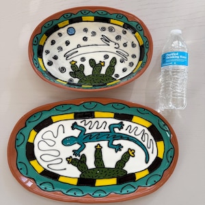 May include: Two oval ceramic serving platters with hand-painted designs. One features a white rabbit and cacti, the other a blue lizard and cacti. Both have brown rims, green and yellow borders, and are on a light grey surface.