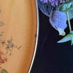 May include: A round plate with a light tan surface featuring a floral pattern of pink roses and green leaves. The plate has a thin metallic gold rim. Beside the plate, there is a cluster of purple flowers with green stems and leaves.