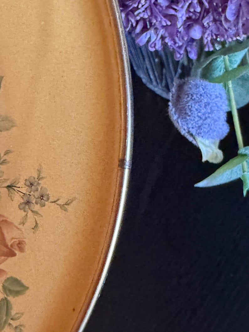 May include: A round plate with a light tan surface featuring a floral pattern of pink roses and green leaves. The plate has a thin metallic gold rim. Beside the plate, there is a cluster of purple flowers with green stems and leaves.