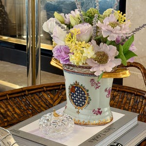 May include: A pale green ceramic vase with gold handles and a decorative crest, filled with a bouquet of lavender, light purple, and yellow flowers. A small, clear glass pumpkin-shaped trinket box with a gold stem sits on a book nearby.