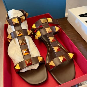 May include: Brown leather sandals with gold pyramid studs. The sandals feature multiple straps and are presented in a red box. These open-toed sandals have a flat sole. A stylish footwear option.
