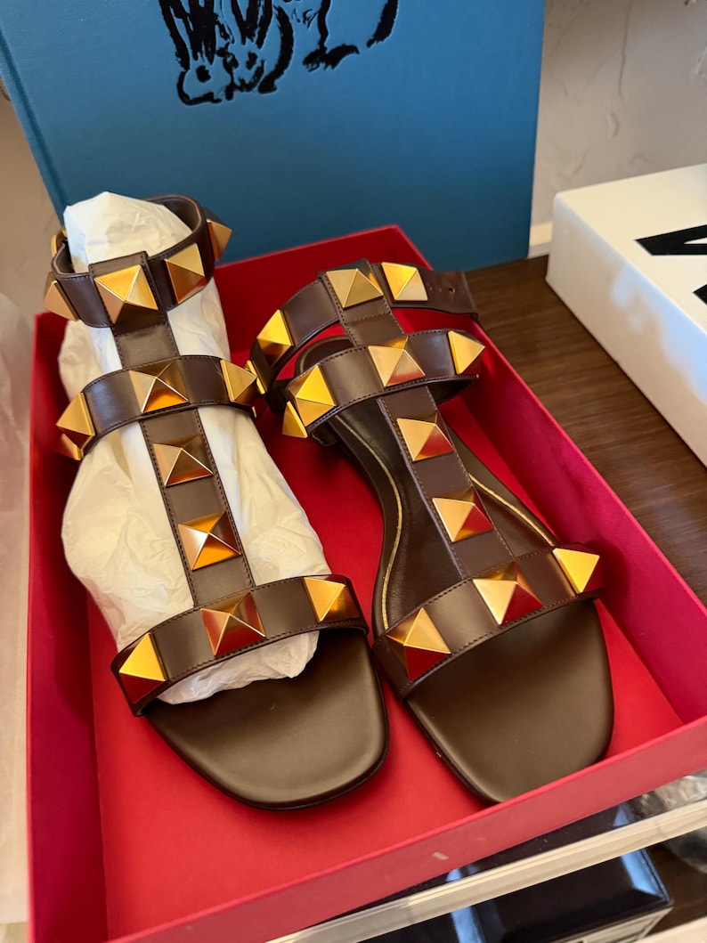 May include: Brown leather sandals with gold pyramid studs. The sandals feature multiple straps and are presented in a red box. These open-toed sandals have a flat sole. A stylish footwear option.