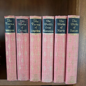 May include: A collection of six antique books with pink covers and gold lettering. The titles include "The Poems of Shakespeare," "The Works of Carroll," and "The Poems of Longfellow." The books are arranged on a wooden shelf.