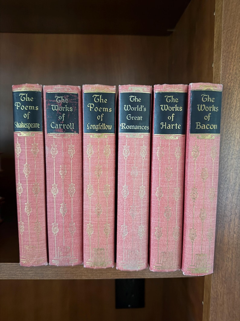May include: A collection of six antique books with pink covers and gold lettering. The titles include "The Poems of Shakespeare," "The Works of Carroll," and "The Poems of Longfellow." The books are arranged on a wooden shelf.