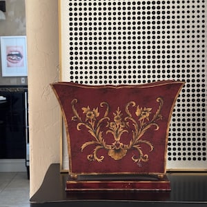 Könnte beinhalten: Ein burgunderfarbener dekorativer Pflanzkübel mit goldenem Blumendesign. Der Pflanzkübel hat eine rechteckige Form mit leicht ausgestelltem Oberteil und passendem Sockel. Das goldene Design zeigt komplizierte Blumenmuster und Verzierungen.