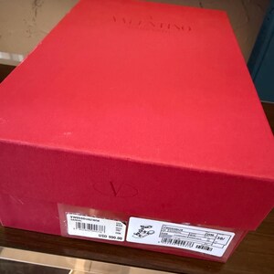 May include: A red rectangular box with the brand name "VALENTINO GARAVANI" embossed on the top. The box has a label with product information, including a style number and the price of USD 990.00.