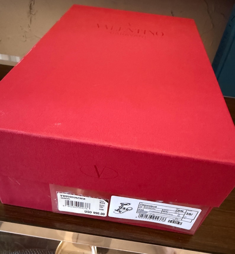 May include: A red rectangular box with the brand name "VALENTINO GARAVANI" embossed on the top. The box has a label with product information, including a style number and the price of USD 990.00.