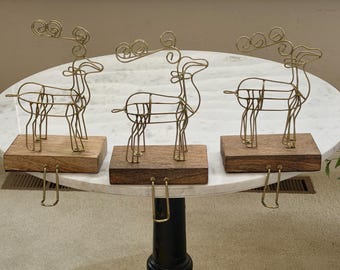 May include: Three decorative reindeer figurines. Each reindeer is crafted from gold-colored wire and mounted on a rectangular wooden base. The reindeer have large, ornate antlers and a hook for hanging stockings. The figurines are displayed on a white marble table.
