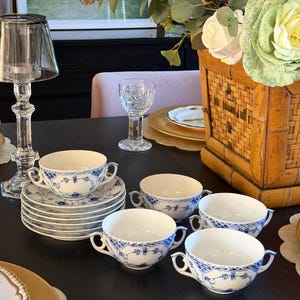 Puede incluir: Un juego de tazas de caldo y platillos de porcelana blanca con un delicado patrón floral azul, bordes intrincados similares al encaje y dos pequeñas asas laterales. Las tazas están dispuestas en una pila e individualmente.