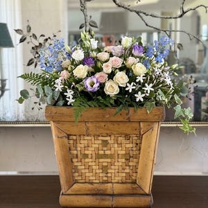 Puede incluir: Una maceta cuadrada de bambú con textura tejida que contiene un arreglo floral de rosas de color rosa claro, blanco y morado, espuelas de caballero azules, flores blancas en forma de estrella y hojas de eucalipto y helechos verdes.