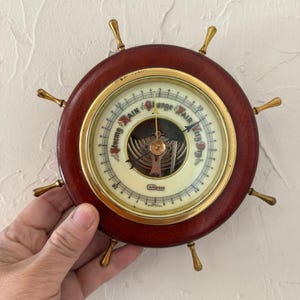 May include: A vintage barometer in the shape of a ship's wheel. The dark wood wheel has brass accents and a cream-colored gauge face. The gauge reads "Storm", "Rain", "Change", "Fair", and "Very Dry". The word "Compass" is printed on the face.