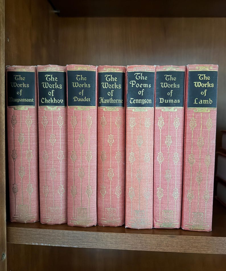 May include: A collection of antique books with pink covers and gold lettering. The spines are black with gold text, and the titles include "The Works of Maupassant," "Chekhov," "Daudet," "Hawthorne," "Tennyson," "Dumas," and "Lamb."