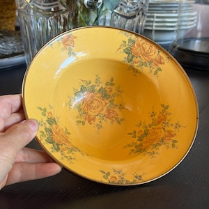 May include: A round, shallow bowl with a golden-yellow base color, featuring a repeating floral pattern of orange and yellow roses with green leaves. The bowl has a thin, metallic gold-toned rim that encircles the entire outer edge of the piece.